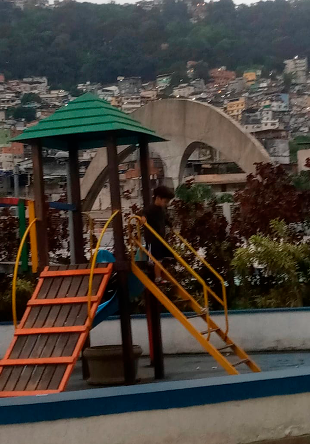 Criança brincando dentro do Complexo Esportivo da Rocinha, tendo ao fundo uma parte da passarela do Oscar Nimeyer, Rocinha e Mata Atlântica. 2022.